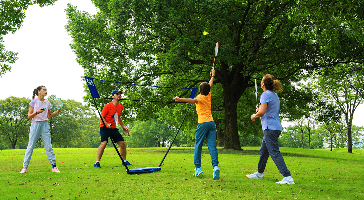 Vier Personen spielen im Park Federball, umgeben von grünem Gras, großen Bäumen und hellem Himmel. Lebendige Farben, Spaß und Bewegung.