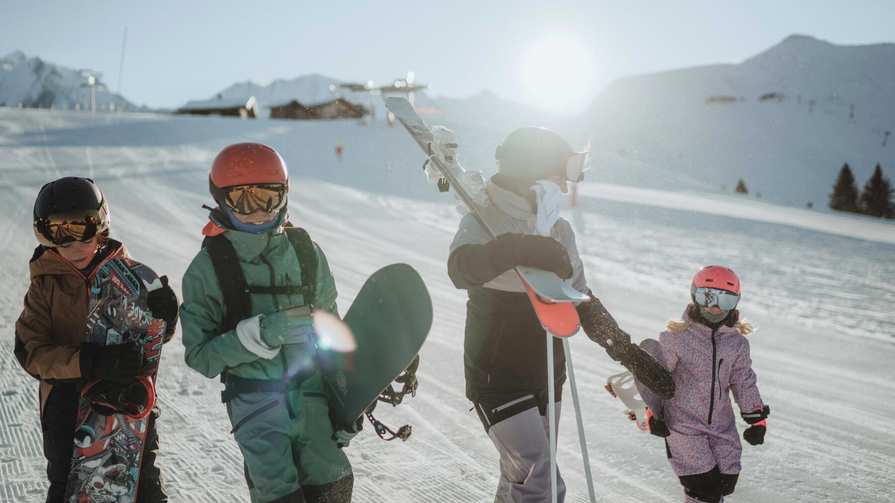 Eine Erwachsener und drei Kinder die mit Snowboards und Ski die Piste runterlaufen.
