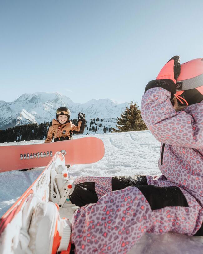 Ein Kind im rosa Skianzug sitzt mit dem Snowboard auf dem Boden. Rechts von ihr ein anderes Kind mit Snowboard, welches ihr zuwinkt.