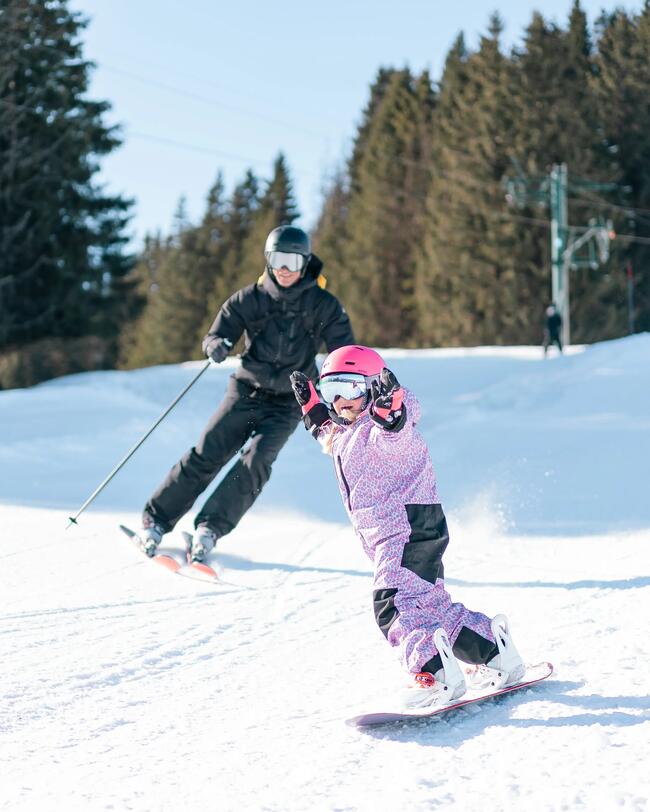 Ein Kind im rosa Skianzug und pinkem Skihelm fährt mit einem Snowboard die Piste runter, dicht gefolgt von einem Erwachsenen in schwarzer Skimontur auf Skiern.