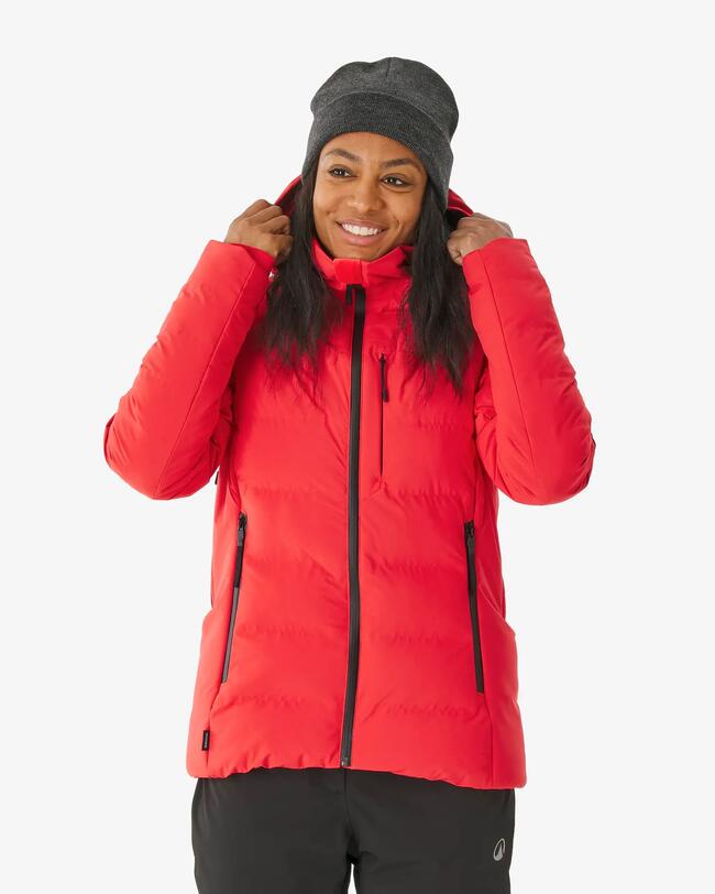 Eine Frau mit roter Jacke und grauer Mütze steht vor einer grauen Leinwand und lächelt.