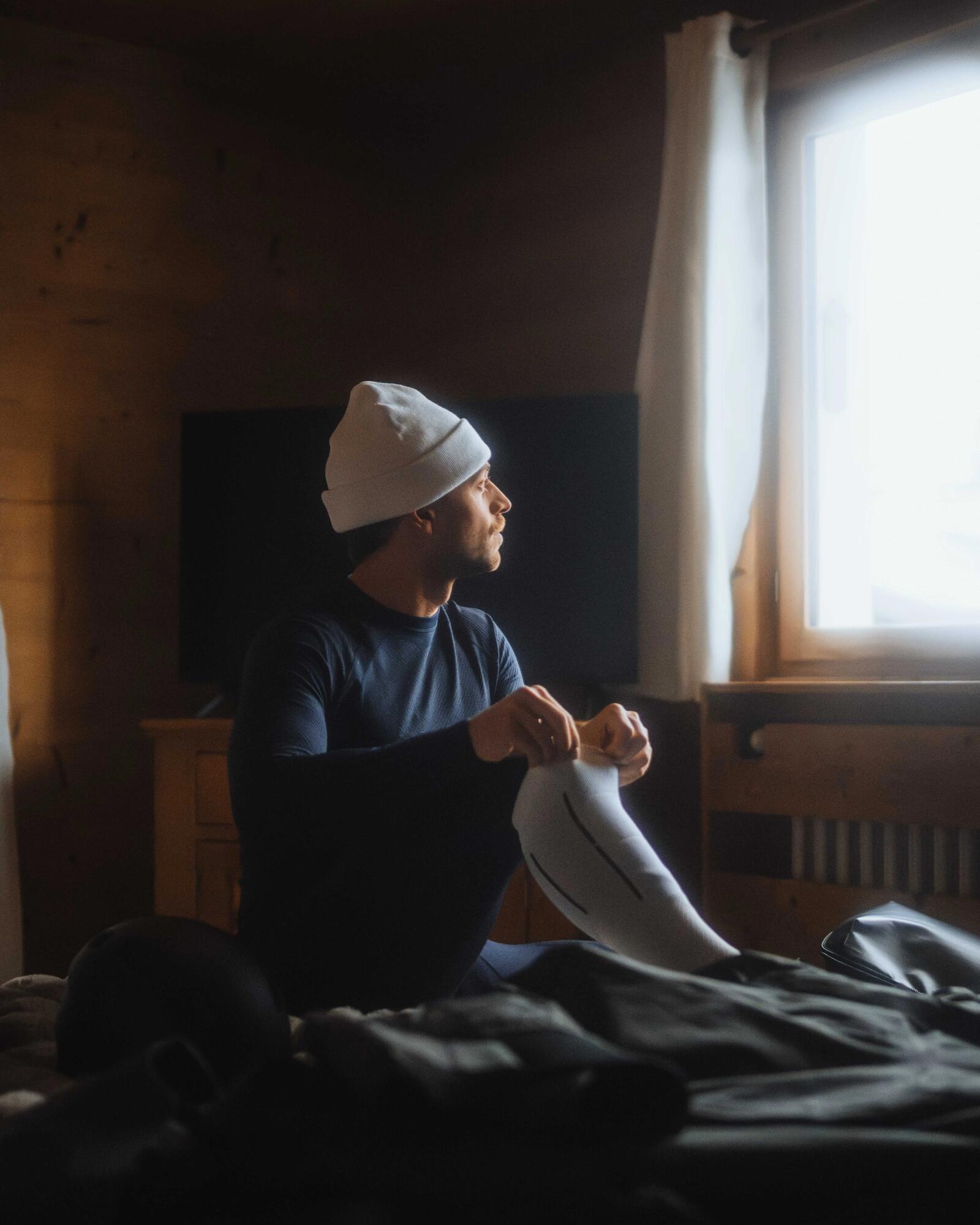 Mann sitzt vor dem Fenster mit Beanie, dabei zieht er sich die Skisocke hoch.