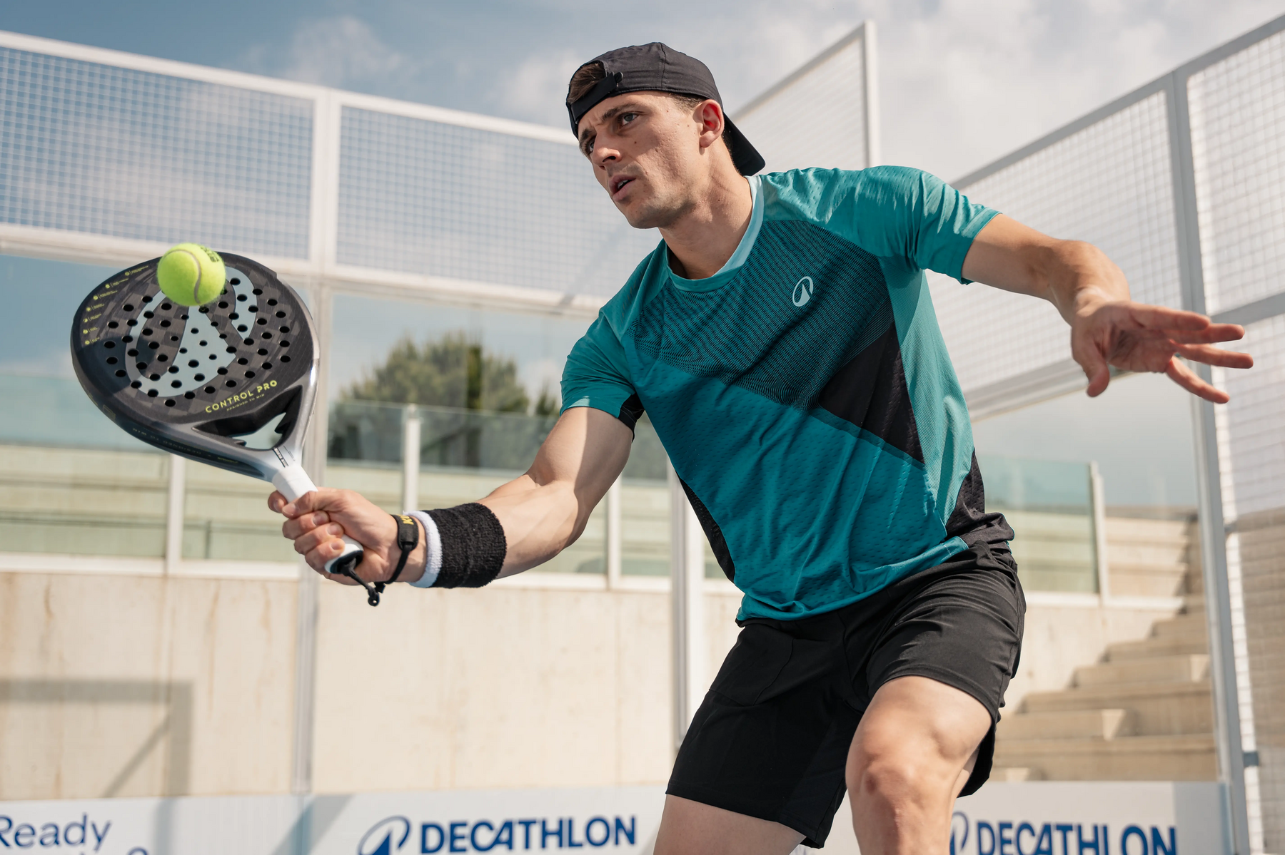Ein junger Padelspieler im tealfarbenen T-Shirt und schwarzer Kappe trifft einen Tennisball mit seinem Padelschläger auf dem Platz.