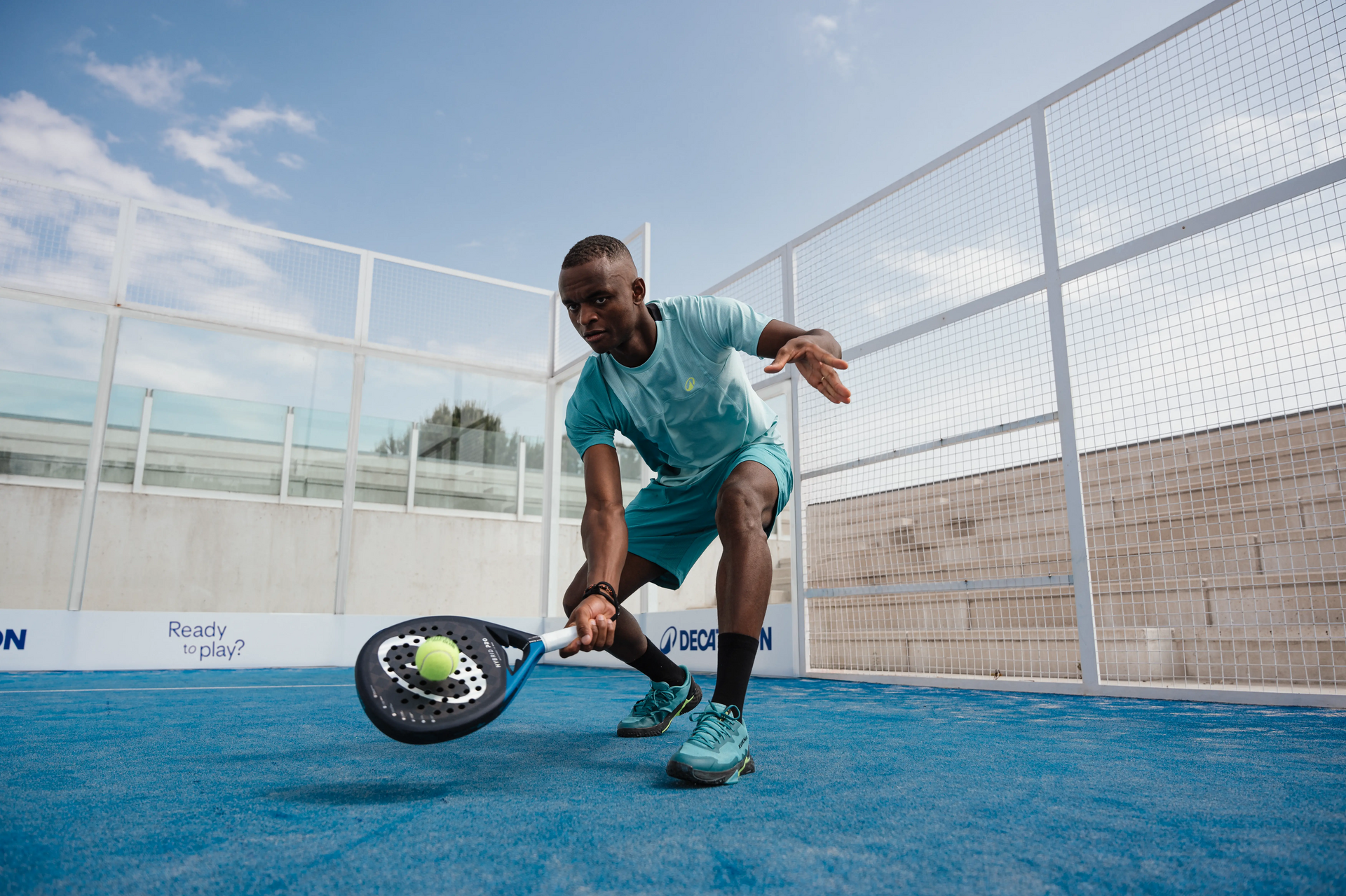 Dieses Bild zeigt einen Padel-Spieler auf einem blauen Padel-Platz, der sich nach vorne beugt und gerade dabei ist, einen gelben Padel-Ball mit seinem Schläger zu treffen.