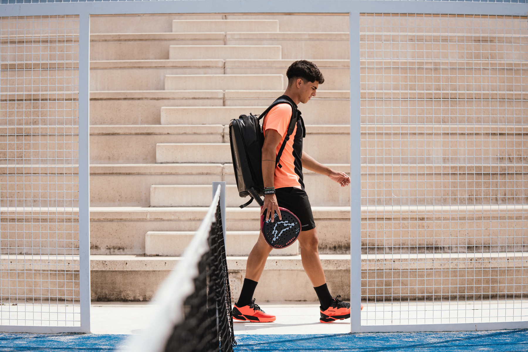 Ein Padelspieler, der einen schwarzen Rucksack trägt, geht mit einem roten und schwarzen Padelschläger in der Hand über den Platz auf eine Tribüne zu.