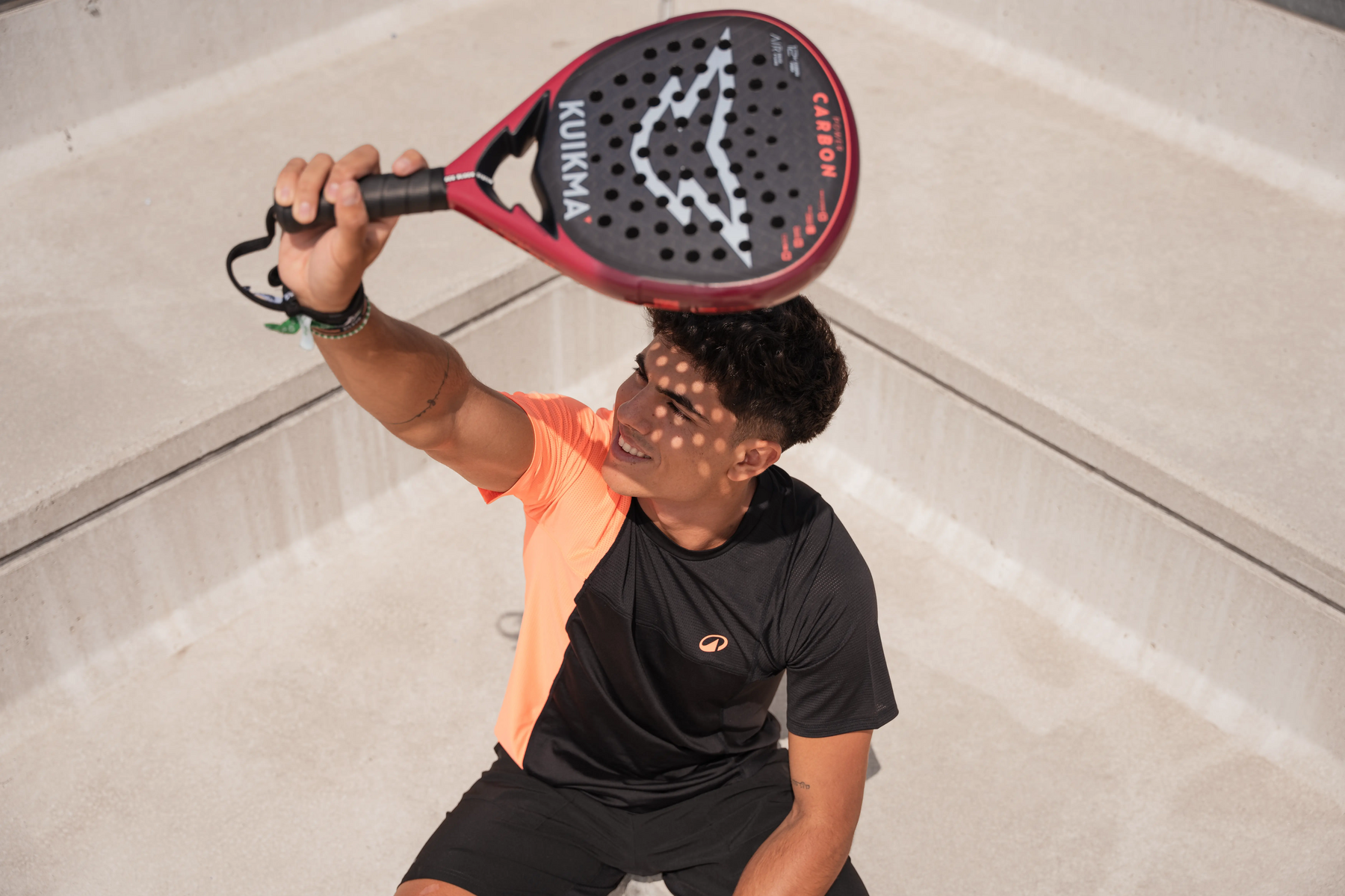 Ein Spieler mit dunklen Locken und einem orange-schwarzen T-Shirt hält ein rotes und schwarzes Padelschläger über seinem Kopf, während er in einem hellen, leeren Becken steht und die Sonne auf sein Gesicht scheint.