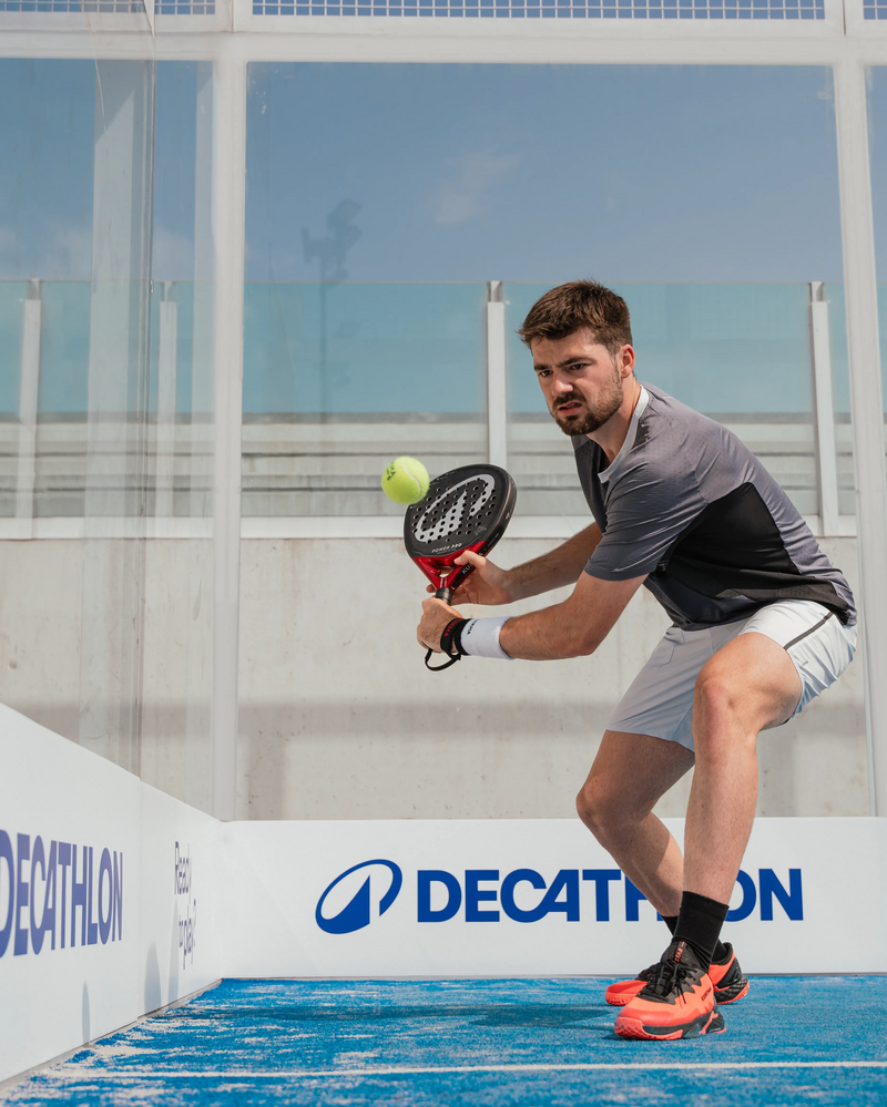 Ein Mann holt mit einem Padelschläger auf einem Outdoor-Padelspielfeld gerade zu einem Schlag auf, während der Ball vor ihm in der Luft schwebt.