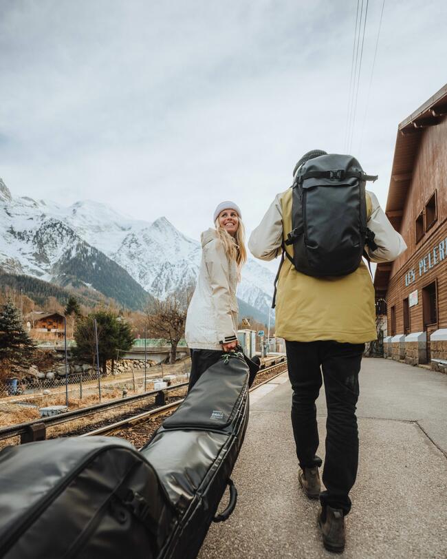 Eine Frau und ein Mann in warmen Klamotten laufen mit Skirucksack, Skitasche und Skischuhtasche an einem Bahngleis entlang.