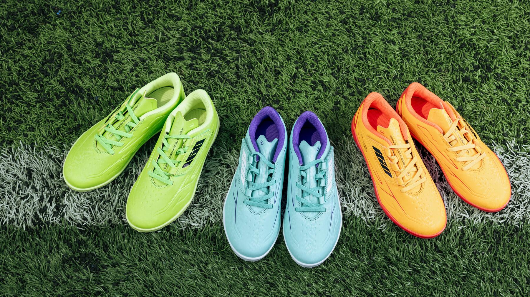Drei Paar bunt gefertigte Fußballschuhe – in Neongrün, Hellblau und Orange – liegen nebeneinander auf einem Kunstrasenplatz.