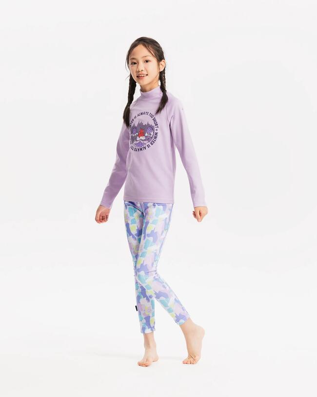 Ein Mädchen mit geflochtenen Zöpfen in hellila Baselayer Top und hellblauer Baselayer Hose steht vor einer grauen Leinwand.
