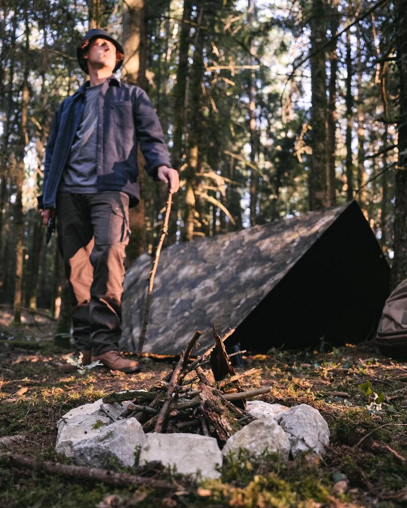 Ein Mann steht im Wald vor seinem aufgebauten Tarp. Davor eine Feuerstelle