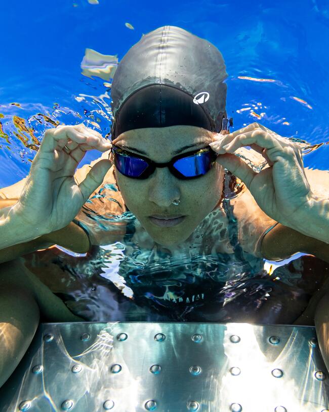 Frau am Beckenrand, Hände an Schwimmbrille, Badekappe, Badeanzug, konzentrierter Blick