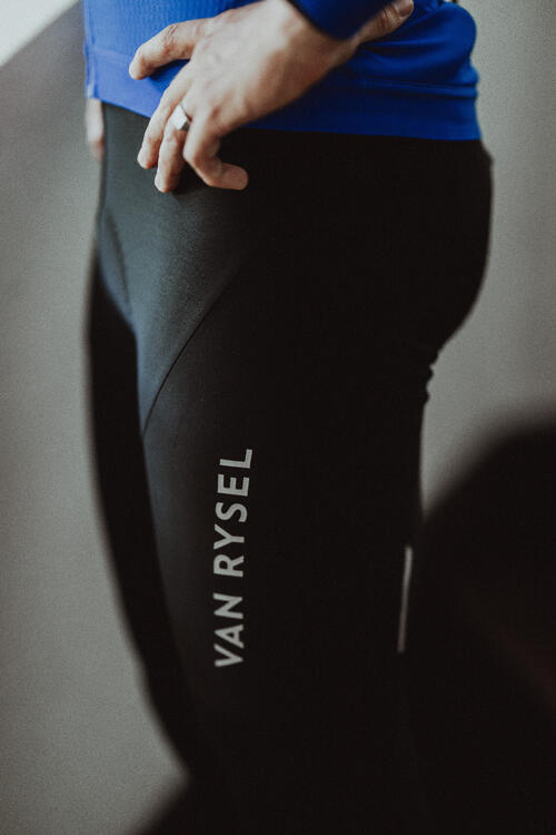 Schwarze, eng anliegende Sporthose mit weißem Van Rysel Schriftzug, blaue Oberbekleidung, matte Textur, Hand auf Hüfte.