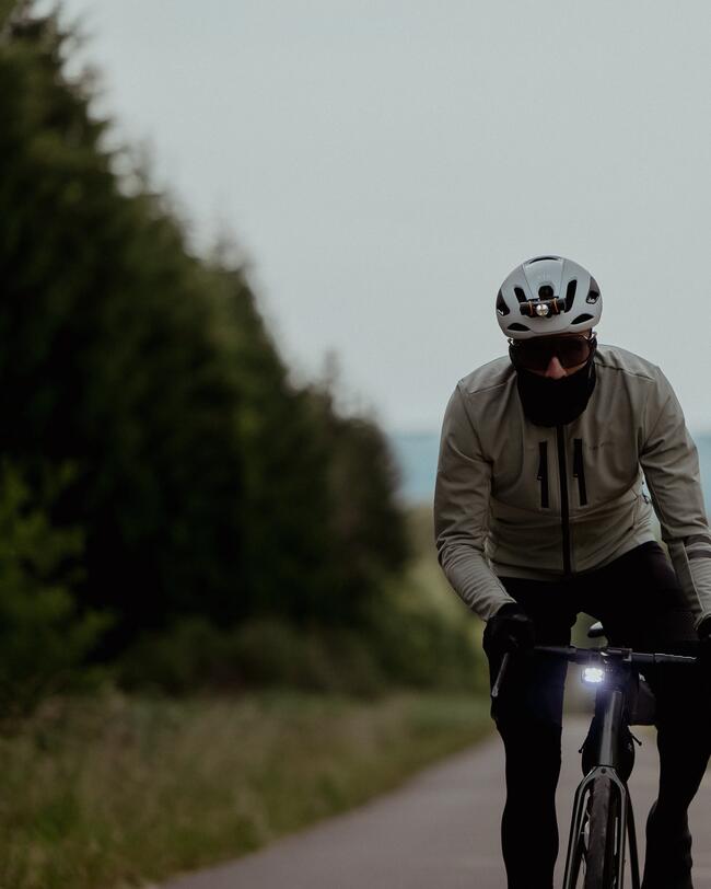 Radfahrer mit grauer Jacke und Helm auf asphaltierter Straße, umgeben von grünen Bäumen, diffuse Lichtstimmung