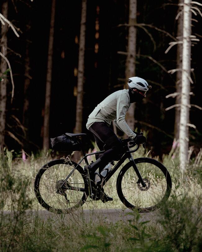Radfahrer mit weißem Helm fährt durch sonnigen Wald; grüne Pflanzen im Vordergrund, Baumstämme im Hintergrund, dunkle Schatten.