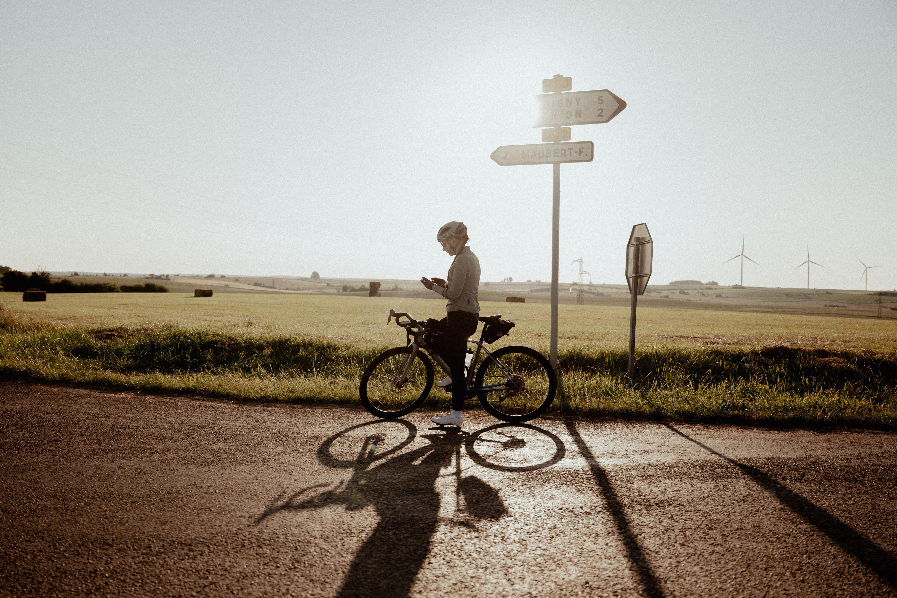 Radfahrer mit Helm steht bei Sonnenlicht auf Landstraße, grüne Felder, Windräder, Straßenschilder im Hintergrund, lange Schatten