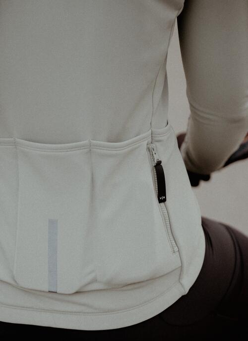Hellgraue Fahrradjacke mit reflektierendem Streifen, schwarze Hose, geflochtener Zopf, hellblaue Fahrradteile sichtbar.
