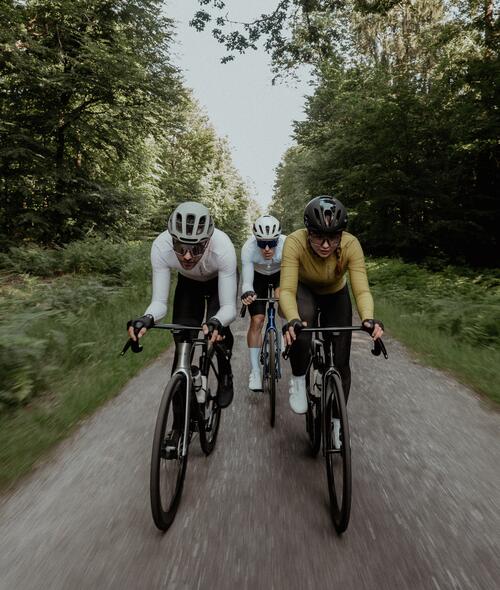 Drei Radfahrer in bunter Sportkleidung fahren auf grauem Weg durch grünen Wald, Sonnenlicht fällt durch das Blätterdach.