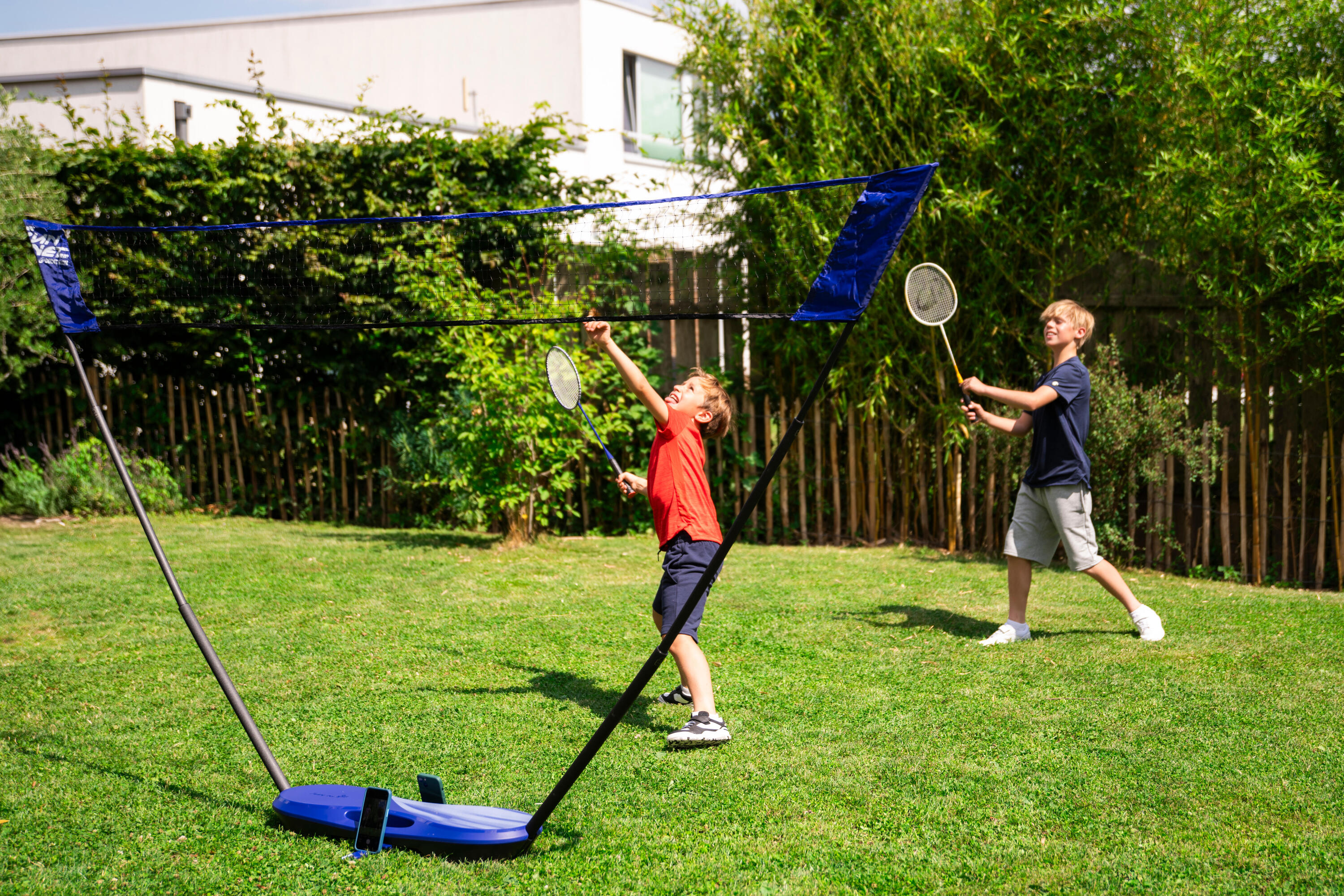 Zwei Jungen spielen mit einem Badmintonset im Garten. Im Vordergrund ist das aufgestellte Netz zu sehen, während im Hintergrund einer der Jungen zum Schlag ausholt.