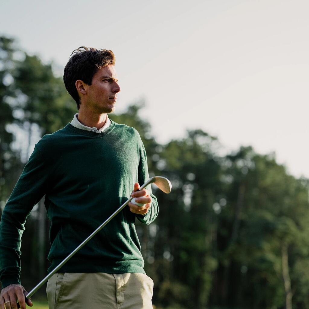Ein Mann im grünen Pullover hält einen Golfschläger und blickt konzentriert über den Golfplatz.