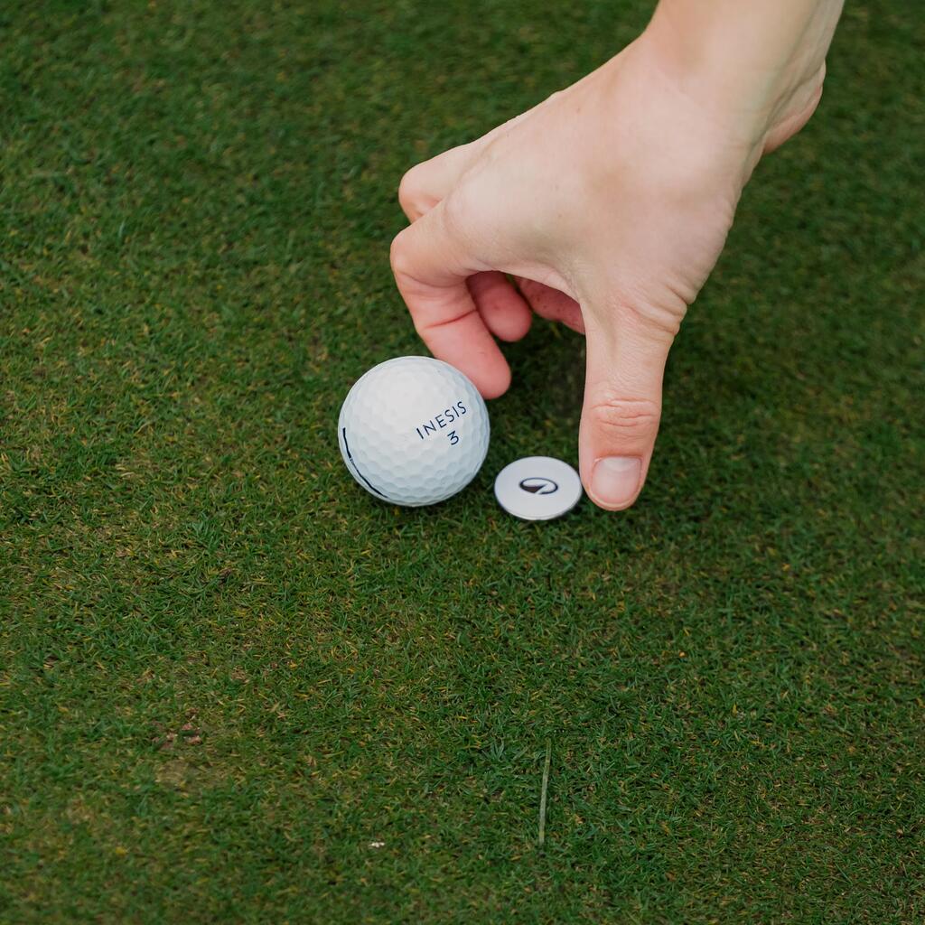 Eine Hand platziert einen kleinen Ballmarker auf dem Rasen direkt neben einem weißen Golfball.