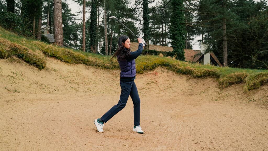 Eine junge Frau schlägt im Sandbunker auf einem Golfplatz Schwung voll den Ball.