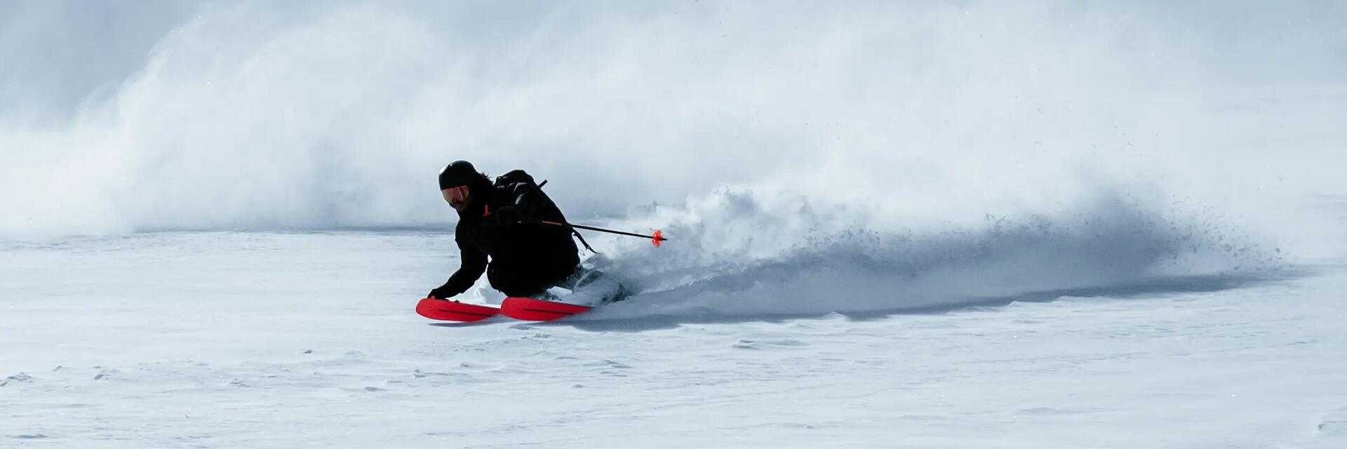 Skifahrerer in schwarzen Skiklamotten und orangenen Skiern auf der Piste.
