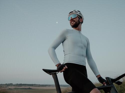 Mann mit weißem Helm und Sonnenbrille, helles Trikot, schwarze Radhose, Fahrrad, blauer Himmel, weiche Lichtstimmung