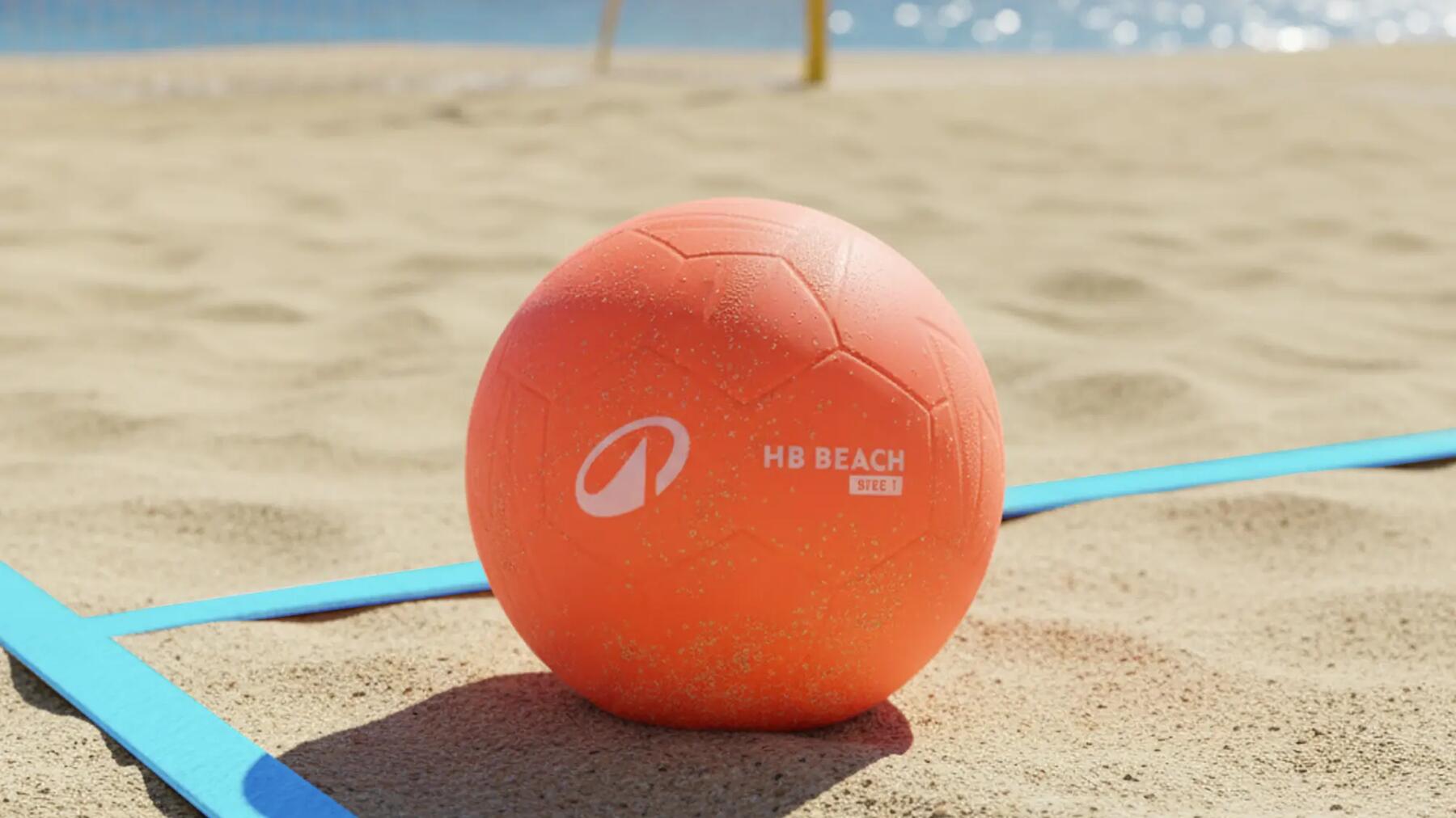 Ein orangefarbener Beachhandball liegt auf dem sandigen Spielfeld eines Beachhandball-Platzes direkt am Meer.