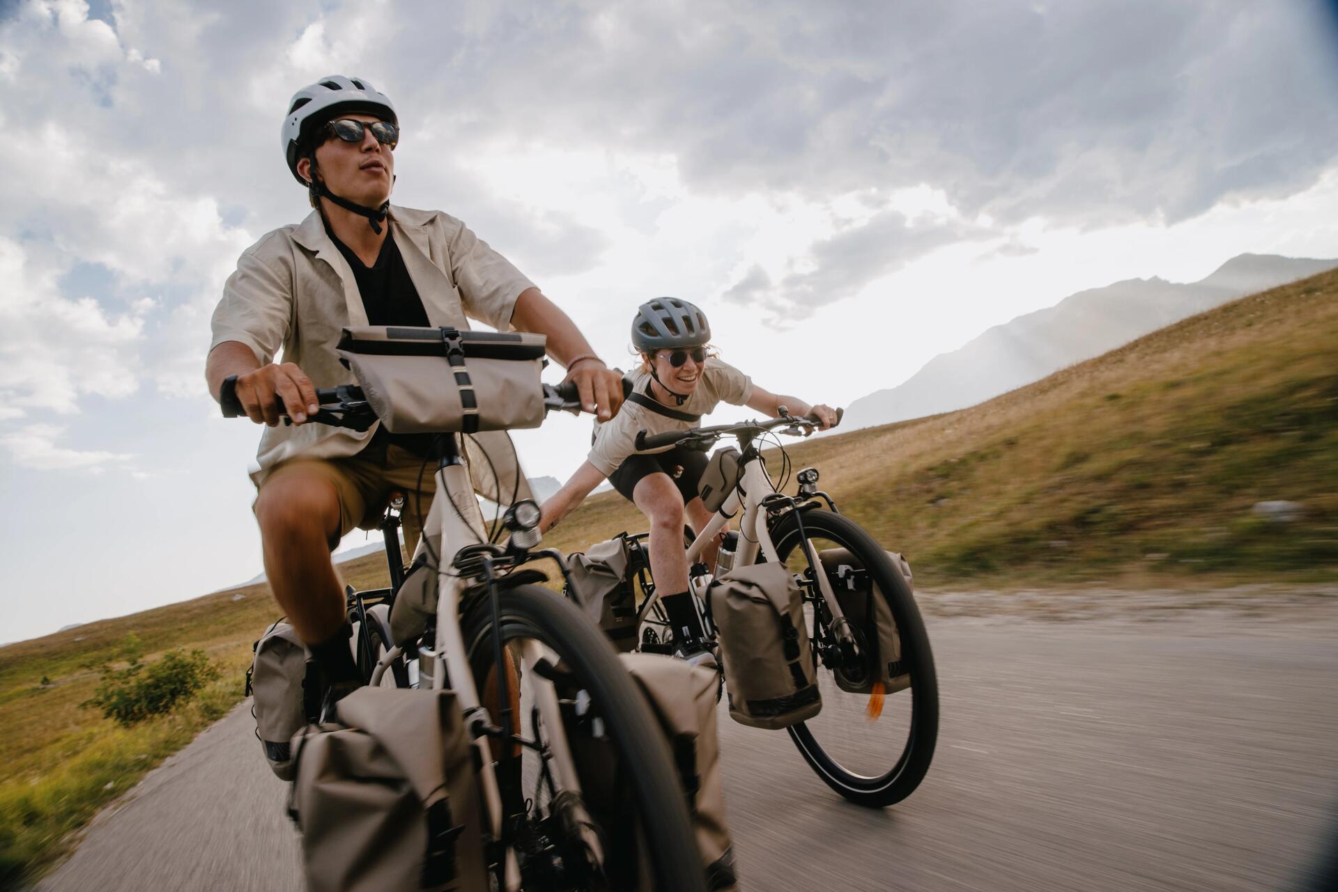 Ein Mann und eine Frau fahren auf vollgepackten Rockrider ADVT 900 Tourenrädern eine asphaltierte Straße entlang. Sie tragen Helme, Sportkleidung und haben Packtaschen vorne und hinten an ihren Rädern befestigt. Die Landschaft ist hügelig mit Wiesen und im Hintergrund sind Berge zu sehen.