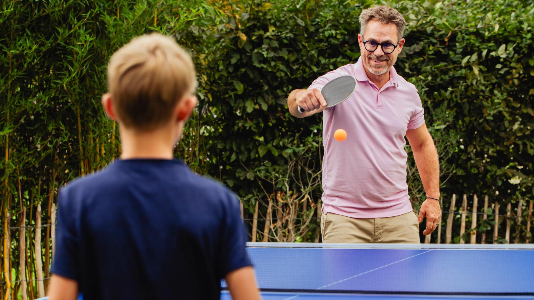 Ein lächelnder Mann im rosa Poloshirt spielt draußen Tischtennis mit einem Jungen, während ein orangefarbener Ball vor ihm fliegt.