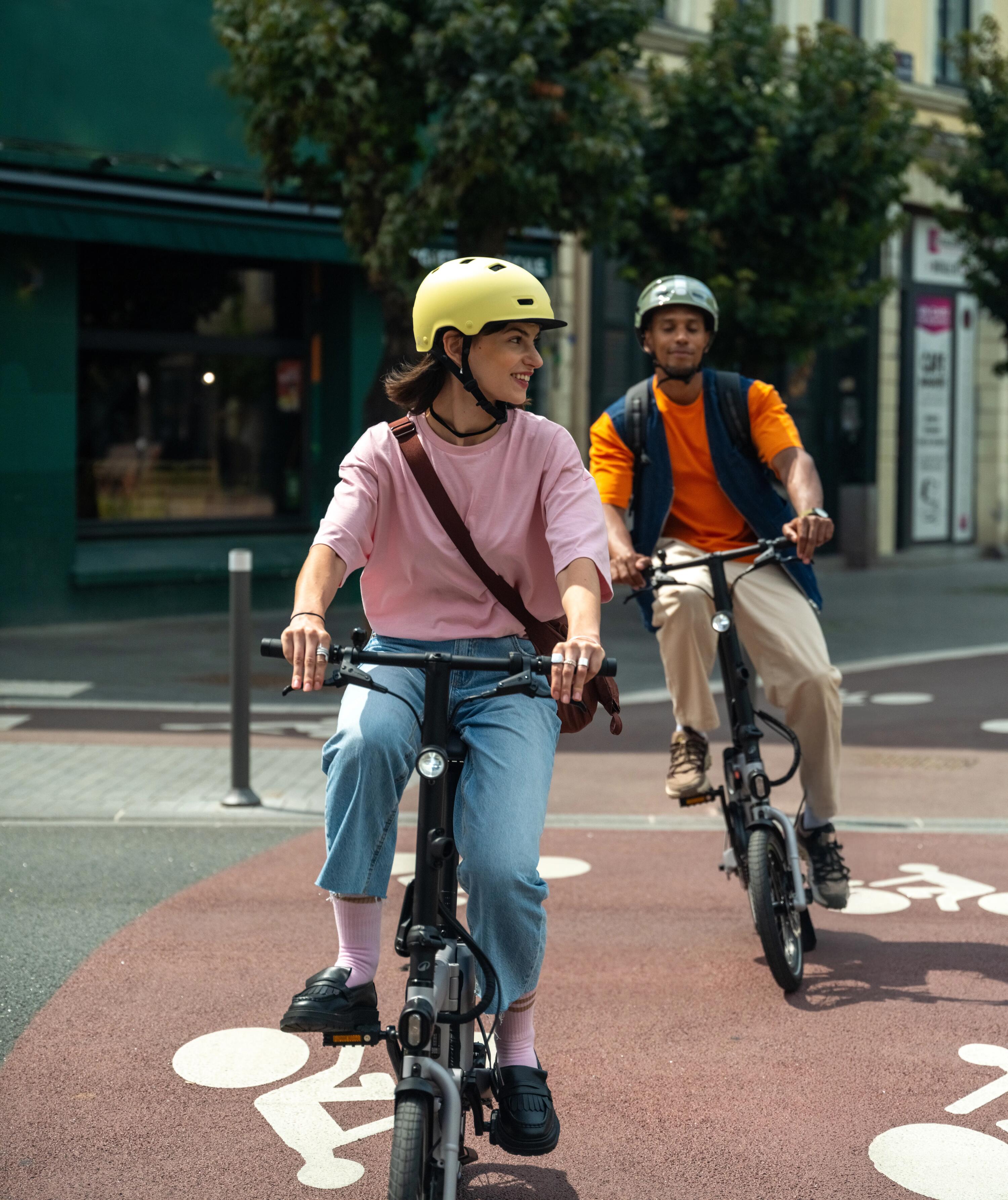 Zwei Personen mit Helmen fahren auf einem roten Radweg auf kompakten E-Falträdern in einer städtischen Umgebung.