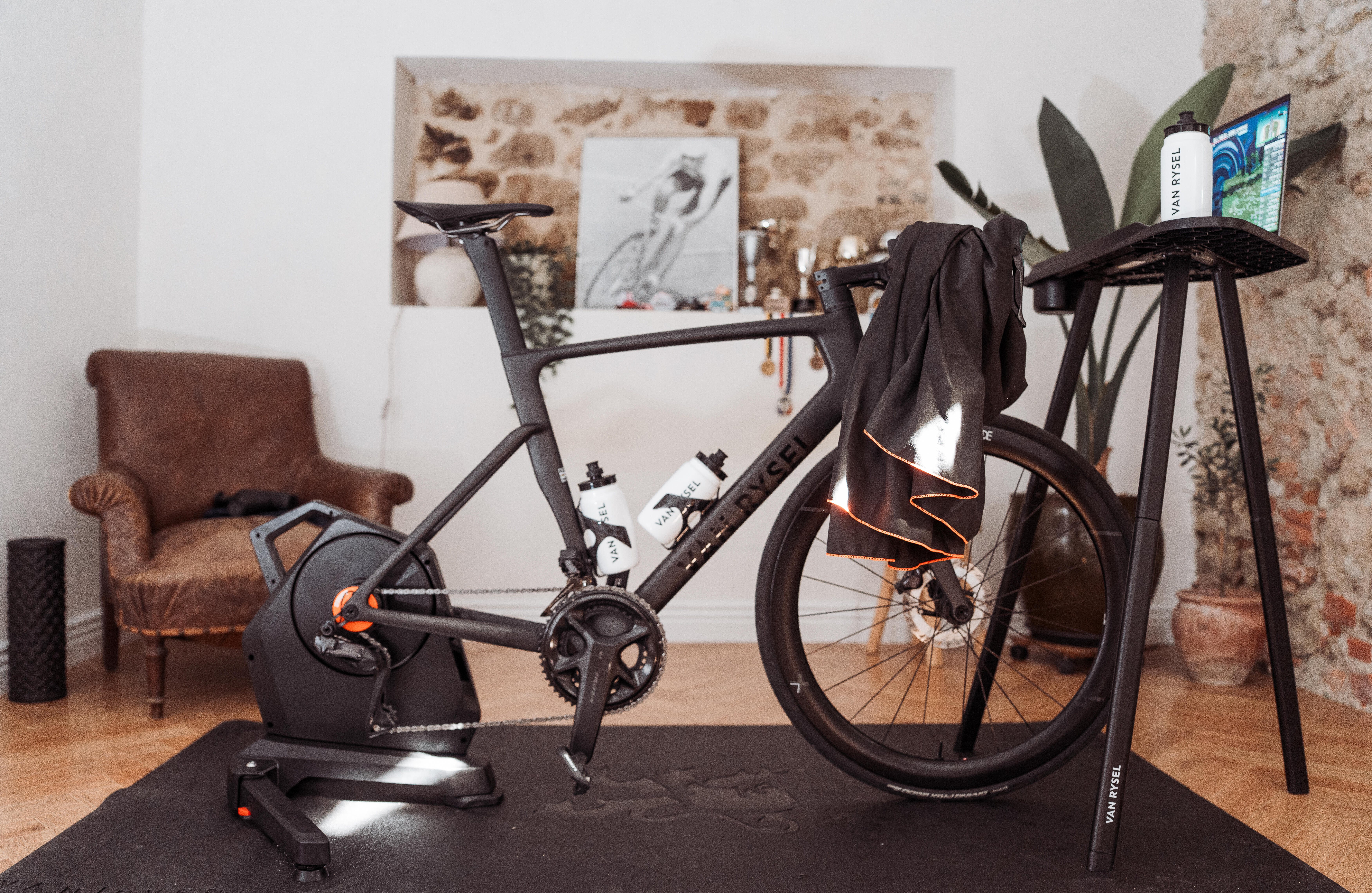In diesem stilvoll eingerichteten Innenraum ist ein matt schwarzes Van Rysel Rennrad auf einem Direct-Drive-Smarttrainer für das Indoor-Training aufgebaut. Der Aufbau umfasst einen Trainingscomputer und ein Handtuch auf einem Ständer, während ein lederner Sessel, Trophäen und eine Fotografie im Hintergrund ein klares Bekenntnis zum Radsport zeigen.