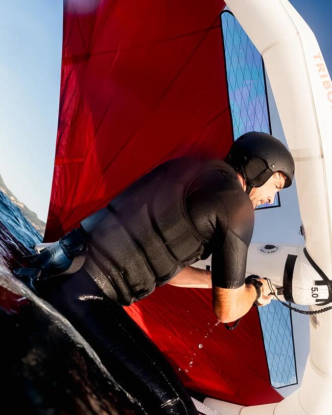 un homme en gilet d'impact assis sur sa planche de wing foil, tenant sa wing tribord rouge