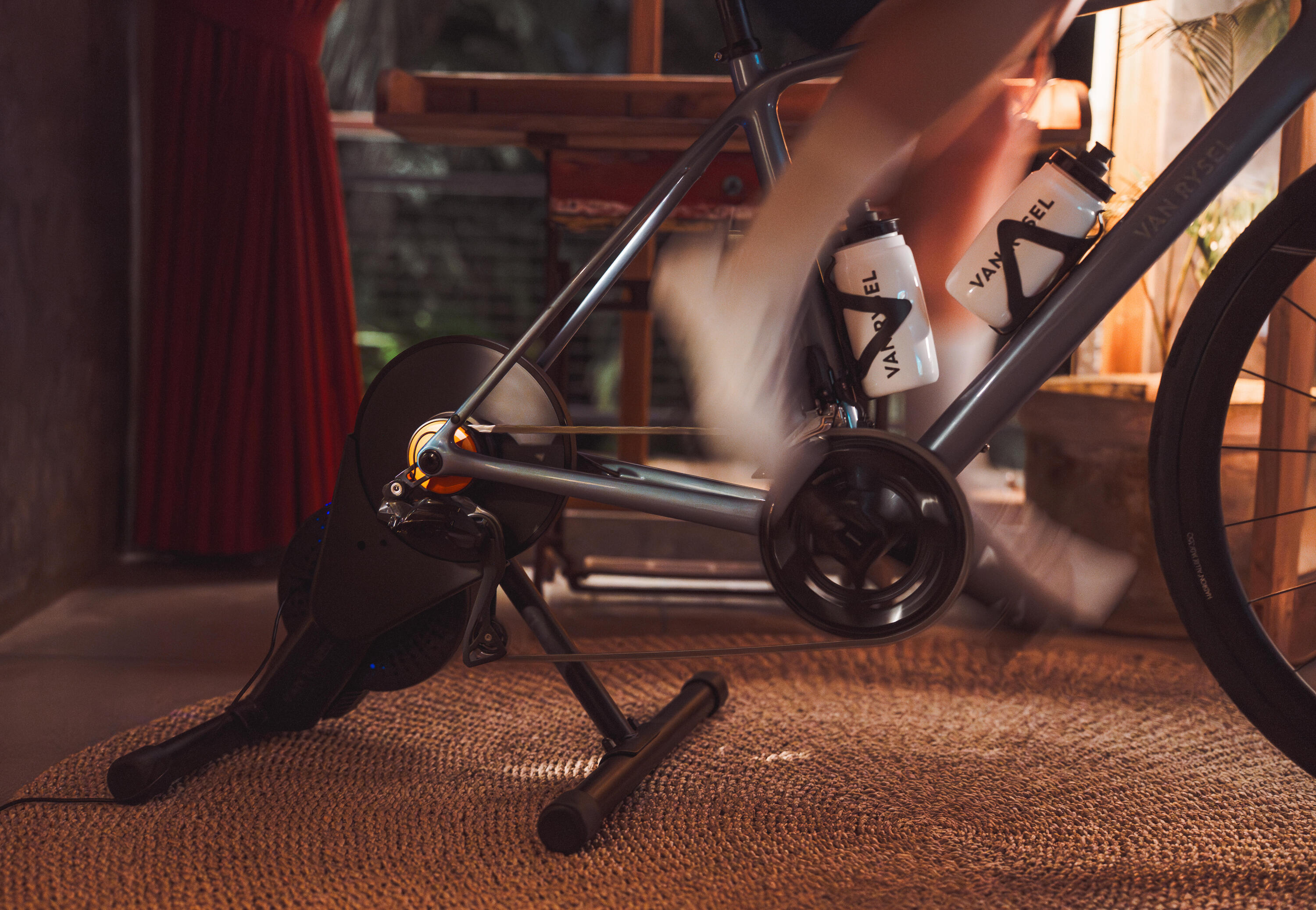 Ein graues Van-Rysel-Rennrad ist für ein Indoor-Workout auf einem Smart-Trainer montiert. Durch eine leichte Bewegungsunschärfe an den Beinen ist zu erkennen, wie die Person in einem gemütlich eingerichteten Raum zügig in die Pedale tritt.
