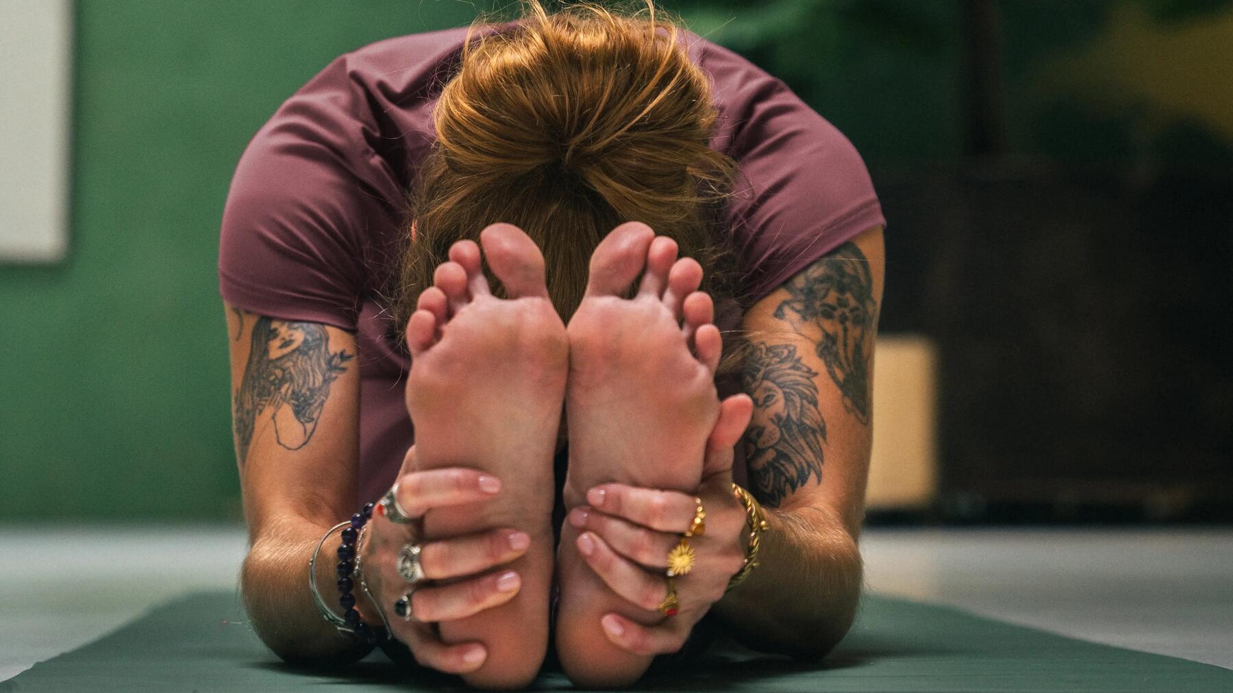 Frau beim Yoga in heller Yoga Kleidung, greift sich in Asana die Füße auf grüner Yogamatte. 