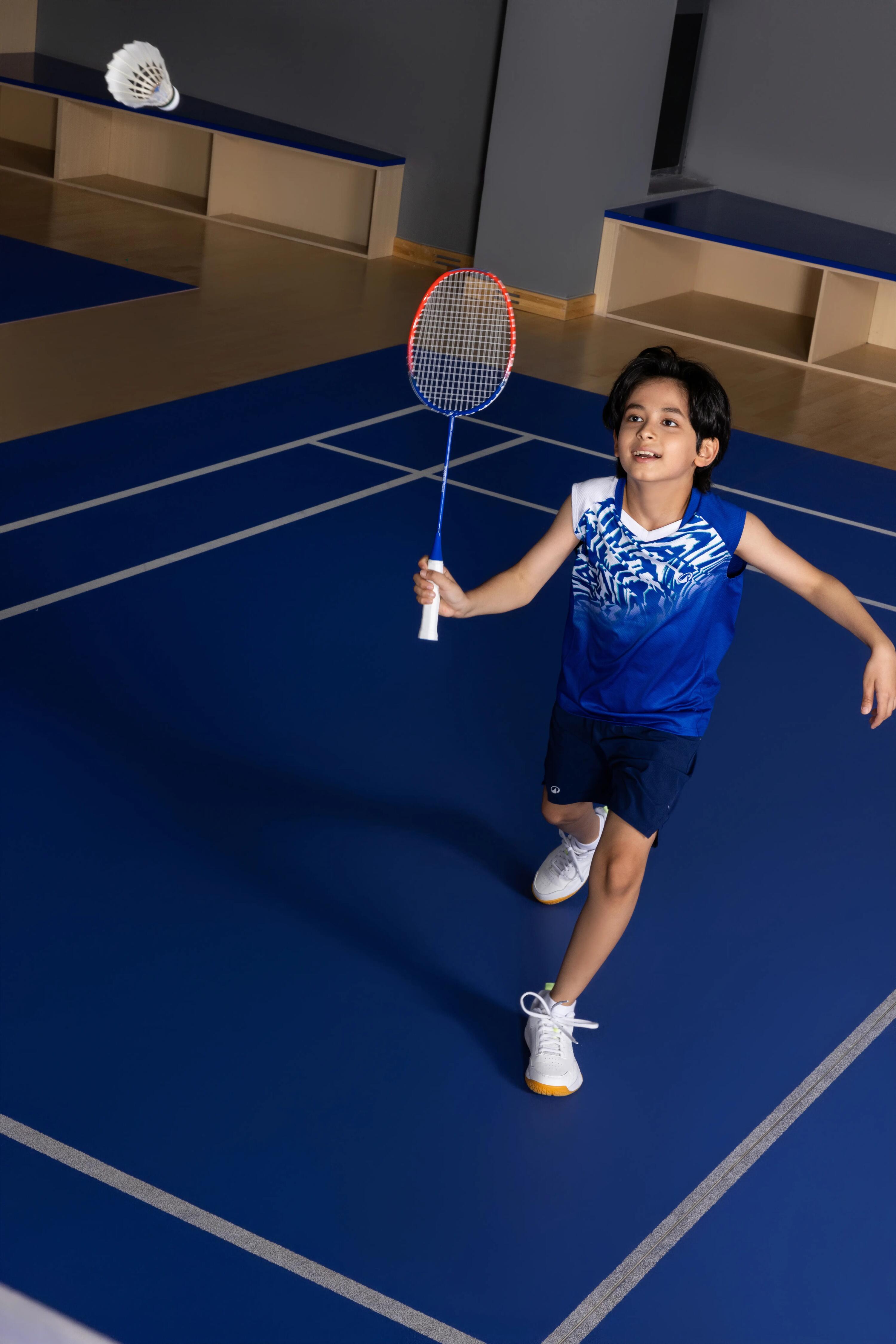 Ein Junge im blauen Trikot spielt Badminton in einer Halle und schaut einem fliegenden Federball entgegen.