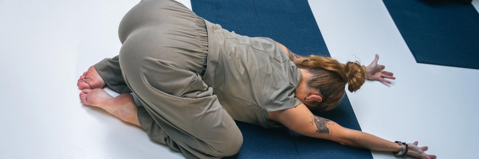 Frau mit Gesicht auf den Boden gerichtet, in grauer Yoga Bekleidung auf dunkelblauer Yogamatte beim Yoga