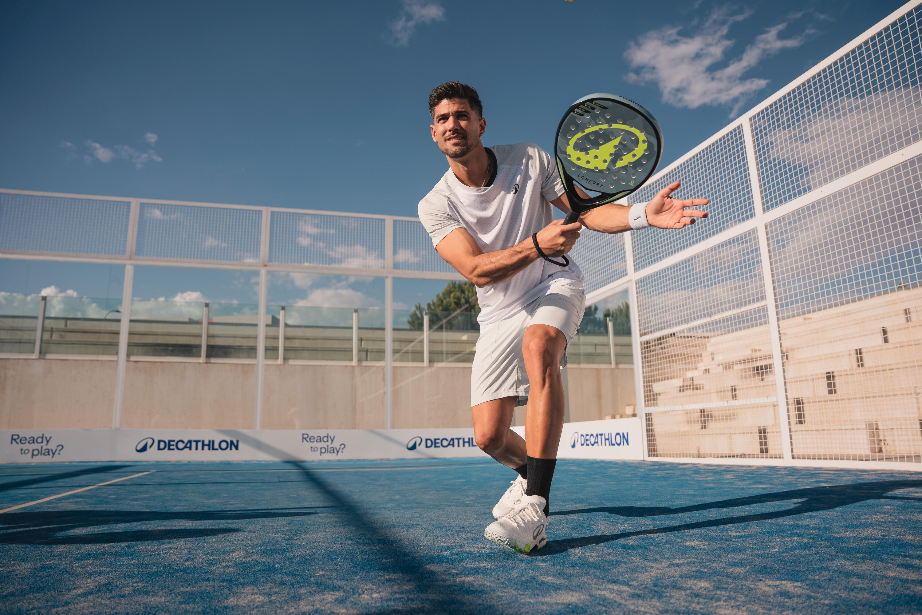 Ein Padel-Spieler in tiefer Hocke schlägt auf blauem Kunstrasen einen gelben Ball knapp über dem Boden. Er trägt komplett weiße Kleidung; im Hintergrund sind Platzumzäunung und Wolkenhimmel zu sehen.