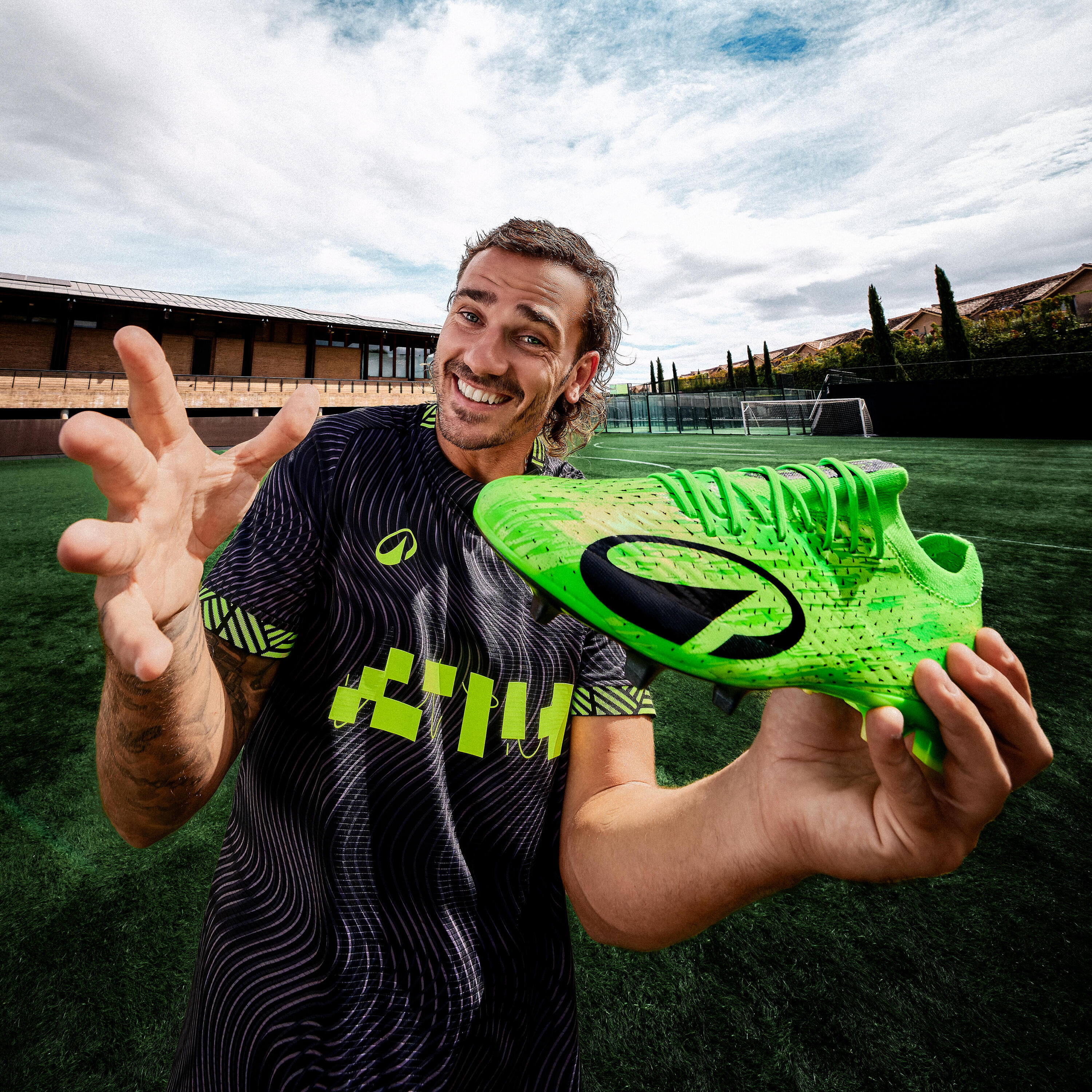 Fußballprofi Antoine Griezmann präsentiert lächelnd den neongrünen Kipsta CLR Fußballschuh auf einem Trainingsplatz.