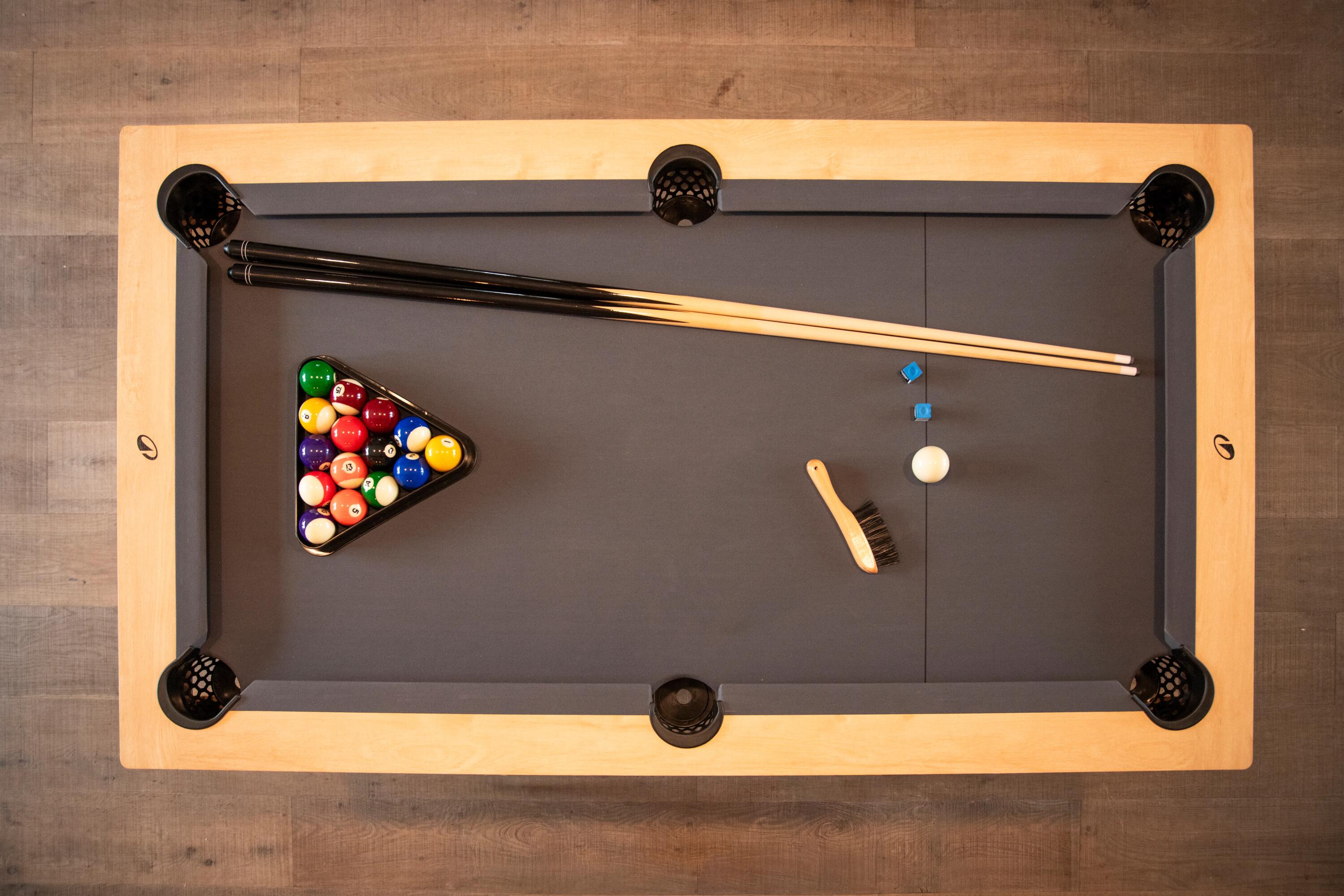 Draufsicht auf einen Billardtisch aus hellem Holz mit grauer Bespannung, aufgebautem Kugel-Dreieck, zwei Cues, weißer Spielkugel, Billardkreide und Tischbürste.