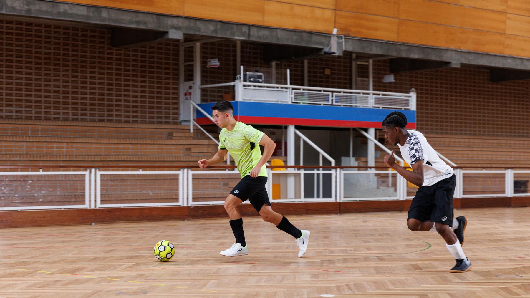 Zwei junge Männer spielen Futsal in einer Sporthalle, während einer von ihnen mit dem Ball am Fuß sprintet und der andere ihn verfolgt.