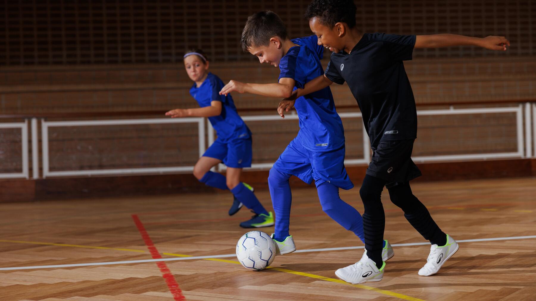 Drei Kinder spielen Futsal in einer Sporthalle, während zwei Jungen im Vordergrund aktiv um den Ball kämpfen.