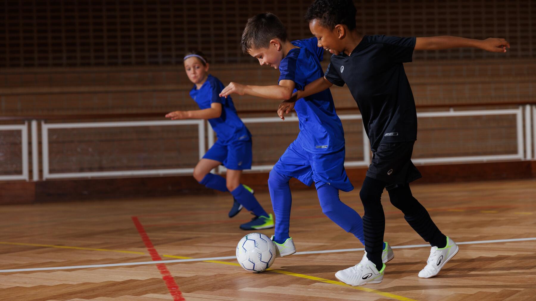 Drei Kinder spielen Futsal in einer Sporthalle, während zwei Jungen im Vordergrund aktiv um den Ball kämpfen.