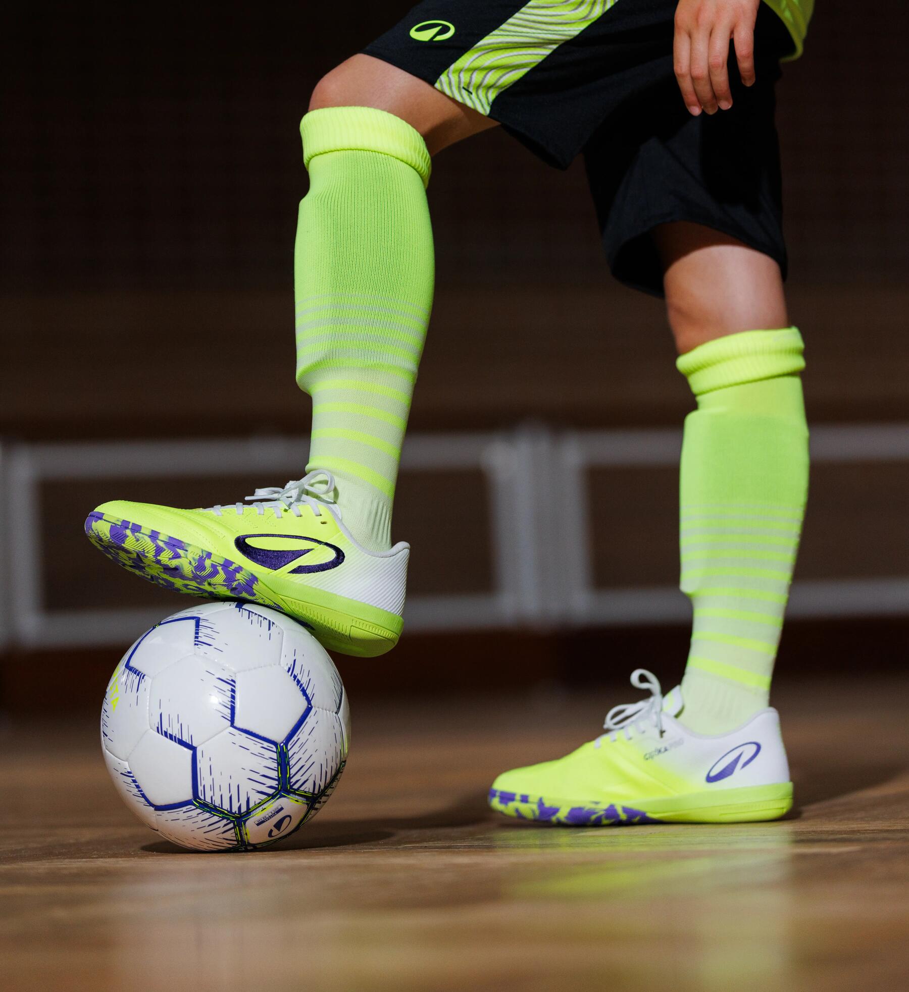 Ein Fußballspieler in neonfarbener Sportbekleidung steht auf einem Hallenboden und hält einen Fuß auf einem weiß-blauen Fußball.