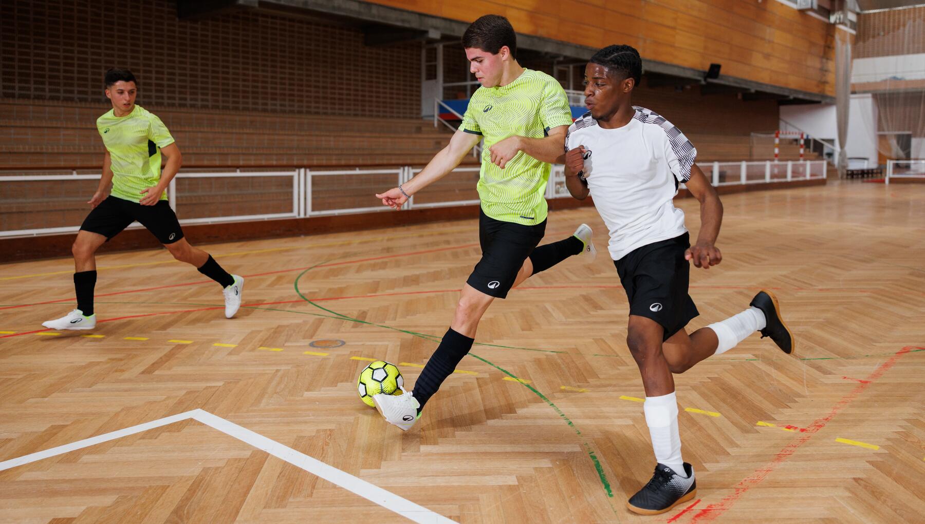 Drei junge Männer spielen Futsal in einer Sporthalle, während einer von ihnen im gelben Trikot den Ball passt und ein Mitspieler ihn verteidigt.