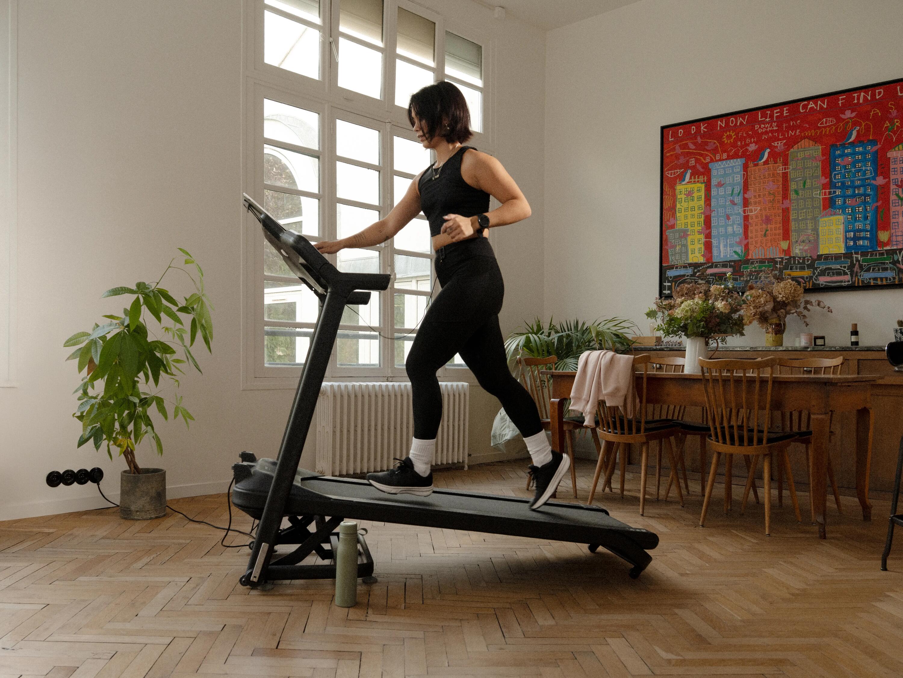 Eine Frau in schwarzer Sportkleidung trainiert auf einem Laufband in einem hellen Zimmer mit Parkettboden.
