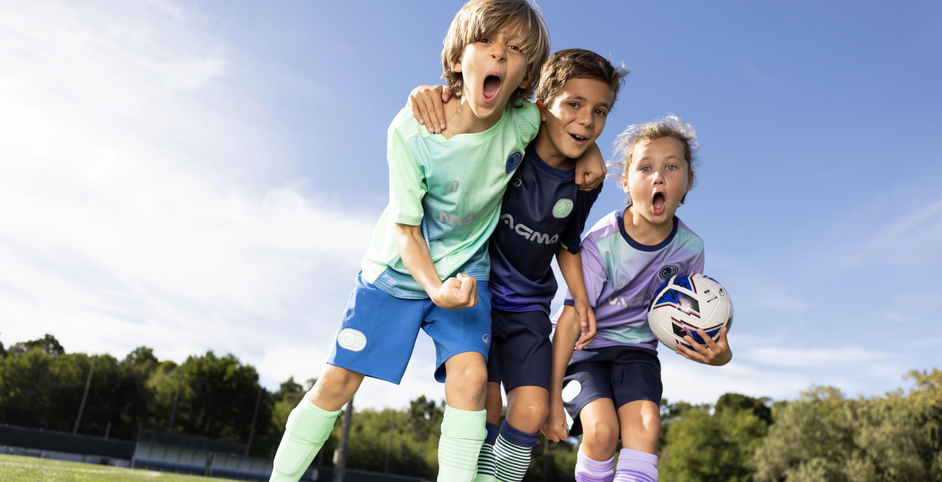 Drei Kinder in bunten Fußballtrikots jubeln ausgelassen und mit weit aufgerissenen Mündern auf einem grünen Sportplatz, während eines von ihnen einen Fußball im Arm hält.