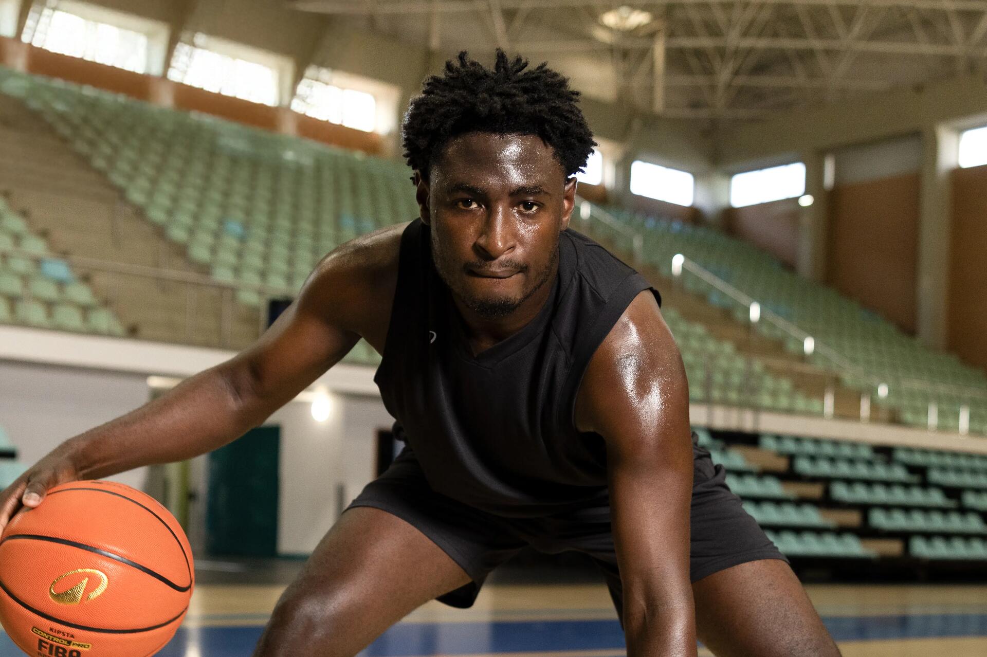 Ein junger, verschwitzter Basketballspieler in schwarzer Sportkleidung dribbelt fokussiert den Ball in einer großen, leeren Sporthalle.