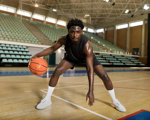 Ein junger Mann dribbelt konzentriert einen Basketball in einer großen, hellen Sporthalle mit leeren Zuschauerrängen im Hintergrund.